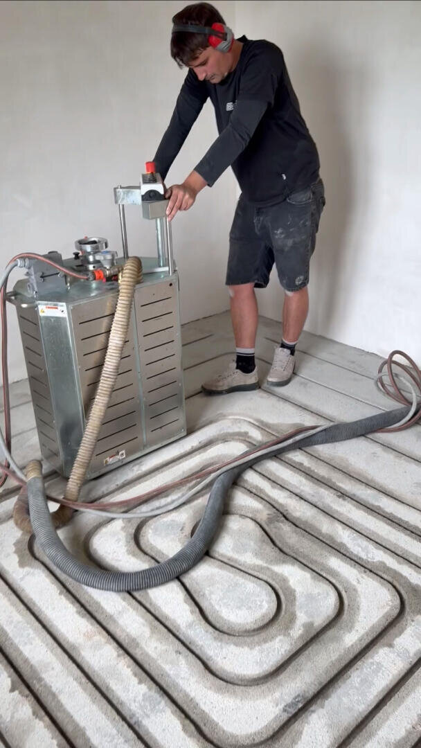 Eine Person bedient eine große Industriemaschine, an der Schläuche befestigt sind, und schneidet Rillen in einen Betonboden für die Installation einer Fußbodenheizung. Der Bediener trägt einen Gehörschutz, ein schwarzes Hemd, kurze Hosen und Turnschuhe.