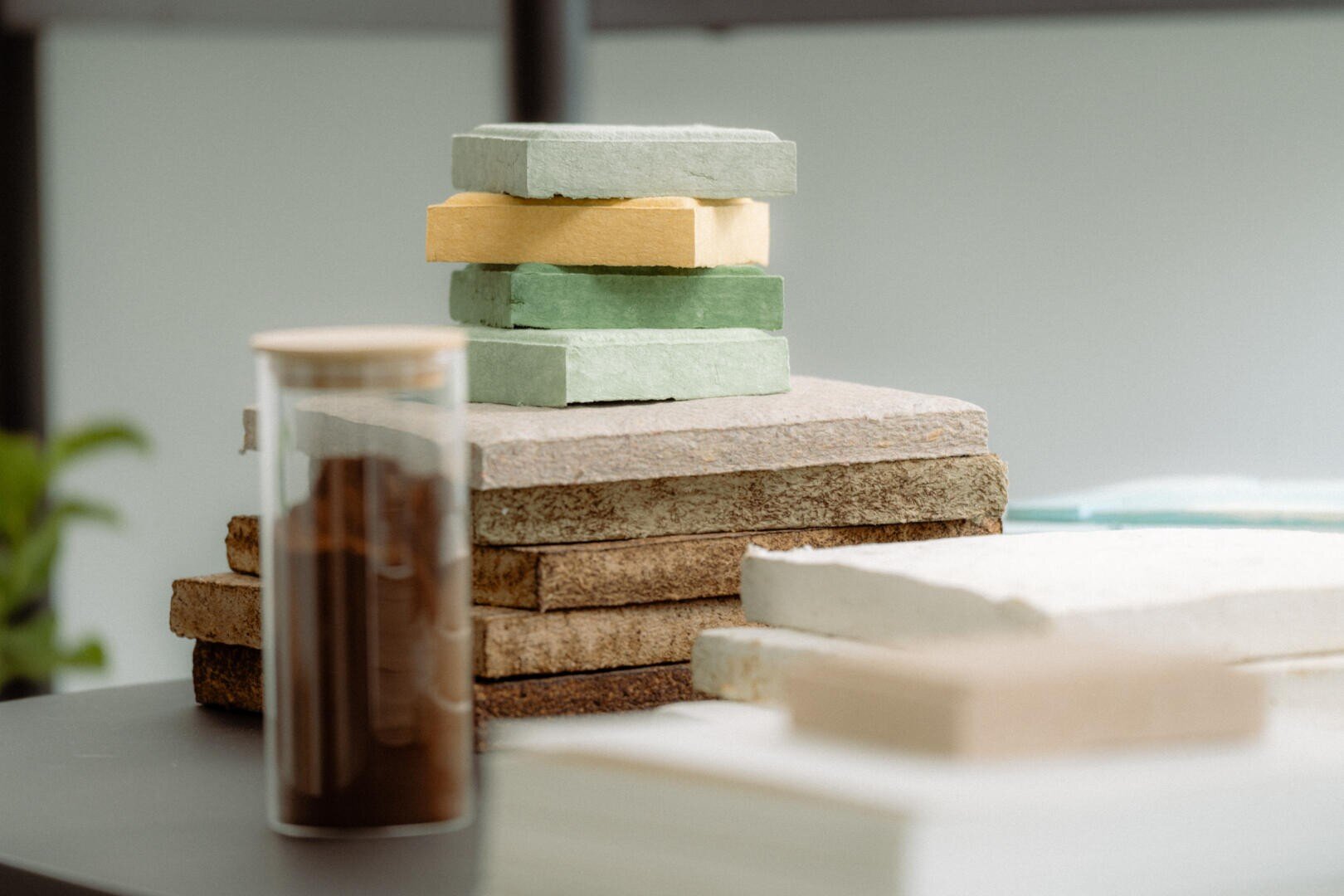 Auf einem Tisch liegt ein Stapel rechteckiger farbiger Ziegelsteine auf größeren, flachen Steinplatten. Daneben steht ein durchsichtiges Glasgefäß, das mit einer dunkelbraunen Substanz gefüllt ist. Der Hintergrund ist unscharf.