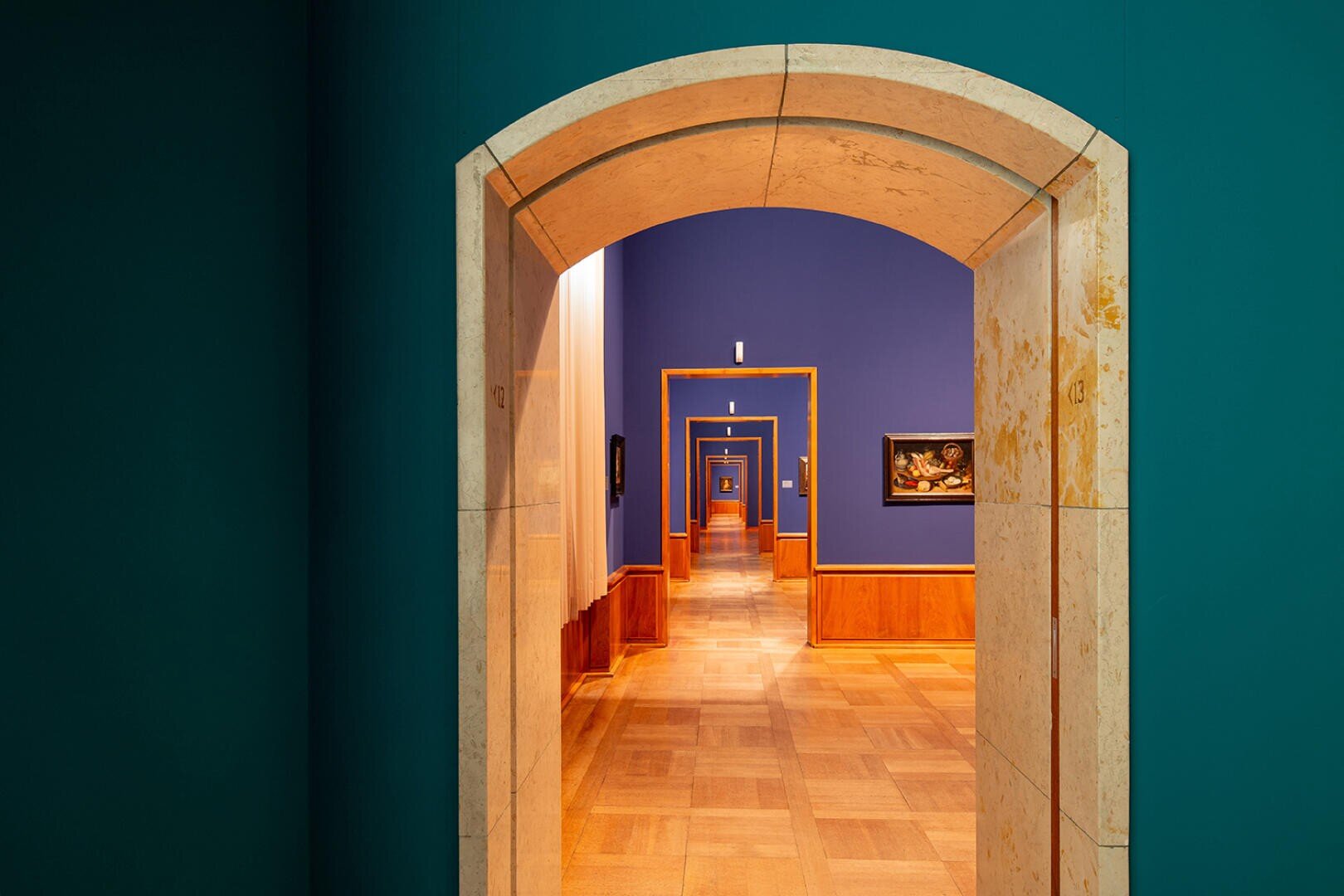 Eine Reihe von gewölbten Türen in einem Museum oder einer Galerie, die in einem langen Korridor mit Holzböden, blaugrünen und blauen Wänden und entlang der Seiten hängenden Gemälden die nächste einrahmen.