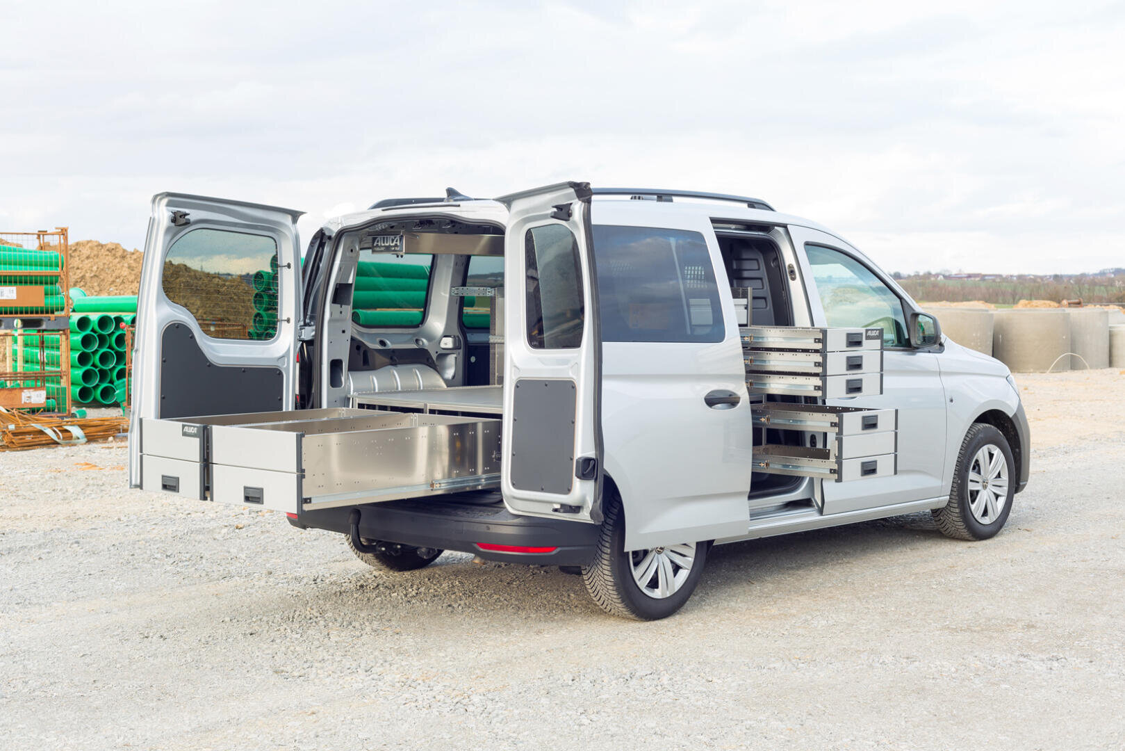 Ein weißer Lieferwagen mit geöffneten Heck- und Seitentüren, der einen aufgeräumten Innenraum mit Metallschubladen und -regalen zeigt, parkt auf einer Schotterfläche auf einer Baustelle mit grünen Rohren und Erdhaufen im Hintergrund.