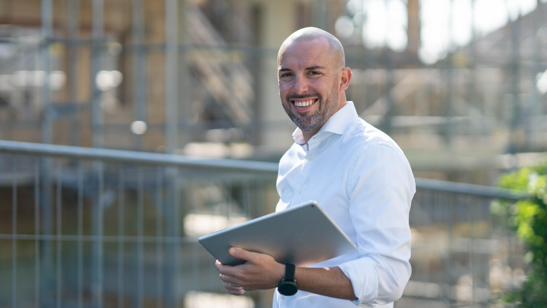 Ein Mann in einem weißen Hemd, lächelnd und mit einem Laptop in der Hand, steht im Freien in der Nähe eines Metallzauns und einer Baustelle. Der Hintergrund ist unscharf und deutet auf eine Industrie- oder Bauumgebung hin.