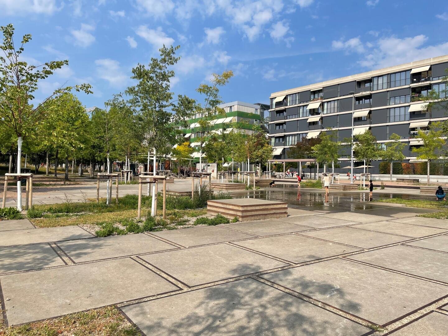 Ein moderner Stadtpark mit jungen Bäumen, Betonwegen und einem Wasserspielbereich, in dem Kinder spielen. Im Hintergrund sind moderne Wohngebäude unter einem teilweise bewölkten Himmel zu sehen.