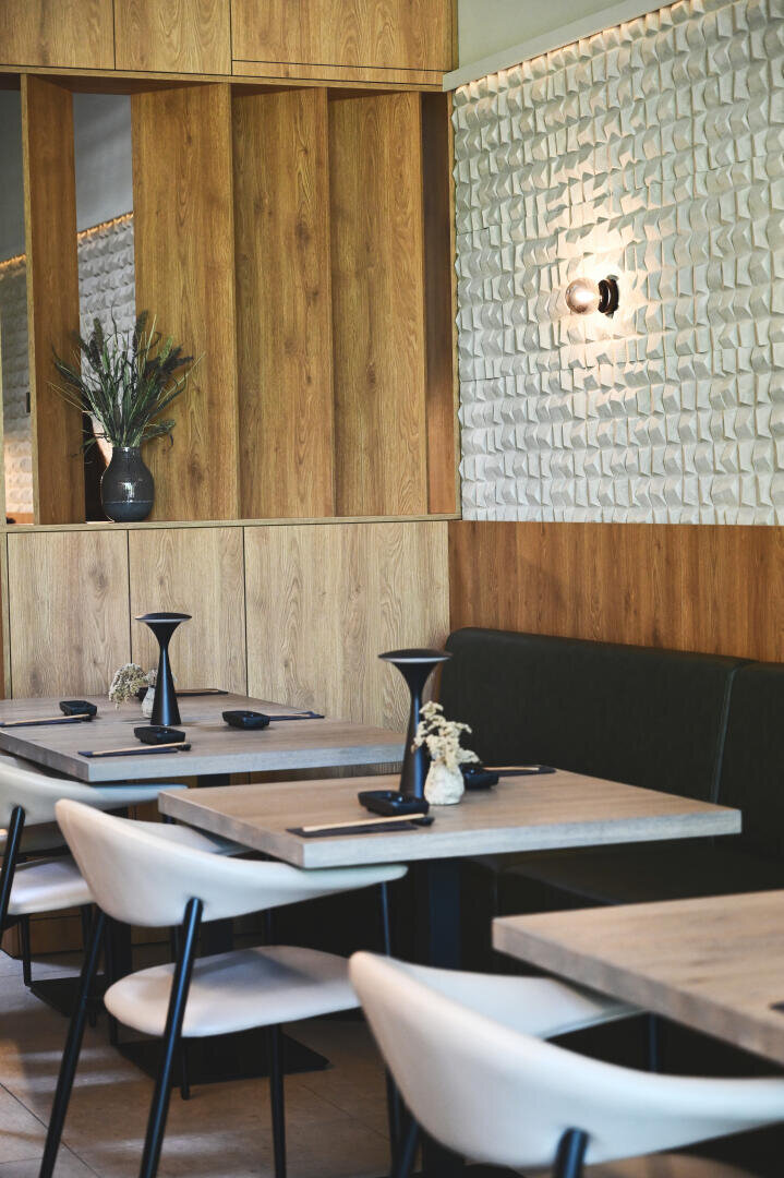 Modernes Restaurantinterieur mit hellen Holztafelwänden, strukturierter weißer Akzentwand, schwarz akzentuierten Tischen, weißen Stühlen und minimalistischem Dekor, darunter eine Vase mit Trockenblumen und Wandleuchten.