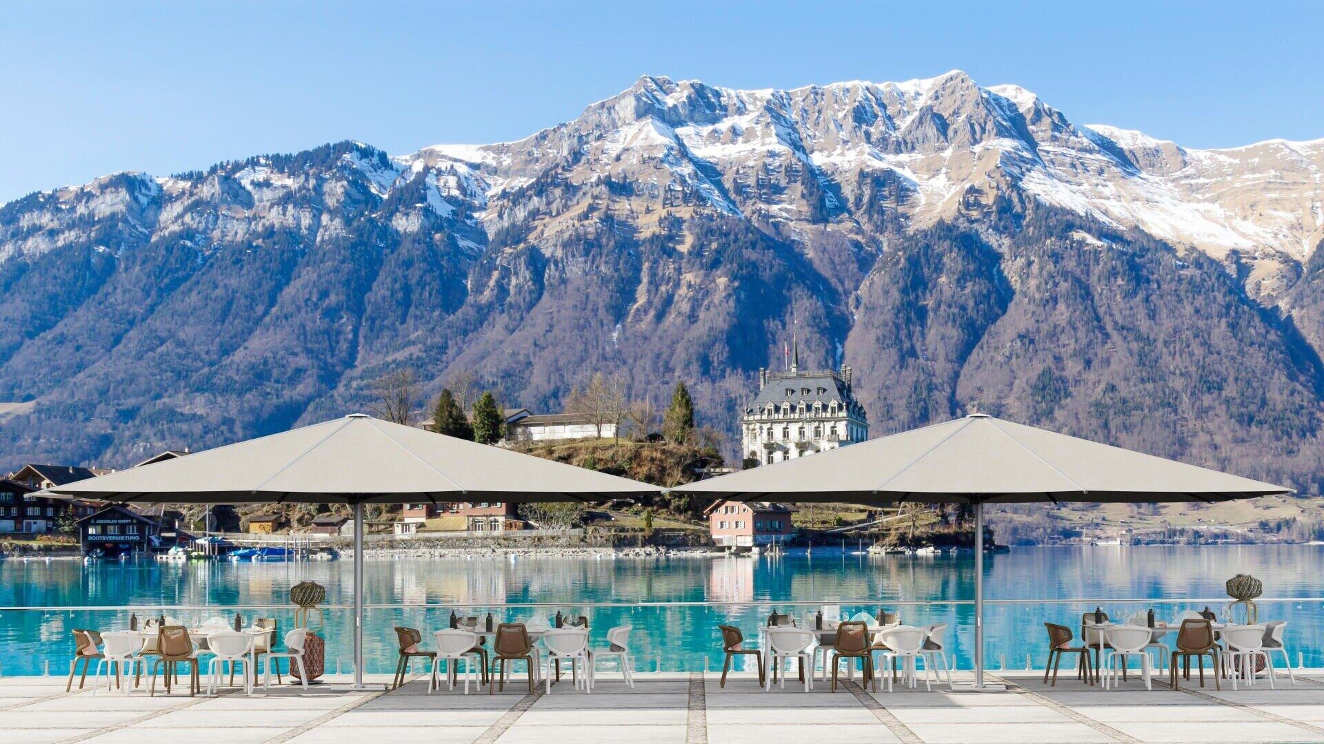 Café im Freien am See mit Tischen und Stühlen unter großen Sonnenschirmen, mit Blick auf einen ruhigen blauen See und schneebedeckte Berge im Hintergrund, mit verstreuten Gebäuden in Ufernähe.