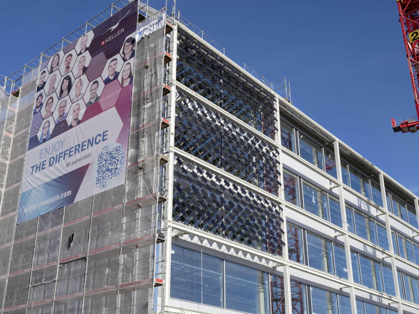 Un immeuble de bureaux moderne en construction affiche une grande bannière avec des visages de personnes, l'inscription Enjoy the Difference et un code QR. Des vitraux bordent la façade ; des échafaudages et une grue sont visibles.