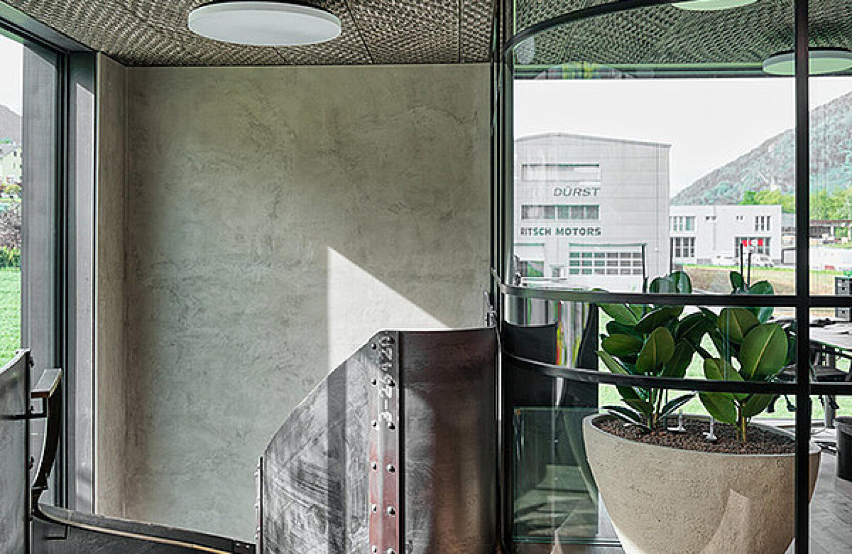Modernes Bürointerieur mit einer großen Topfpflanze an gebogenen Glaswänden, einer Metalltreppe und einem Blick aus dem Fenster auf Industriegebäude und Hügel.