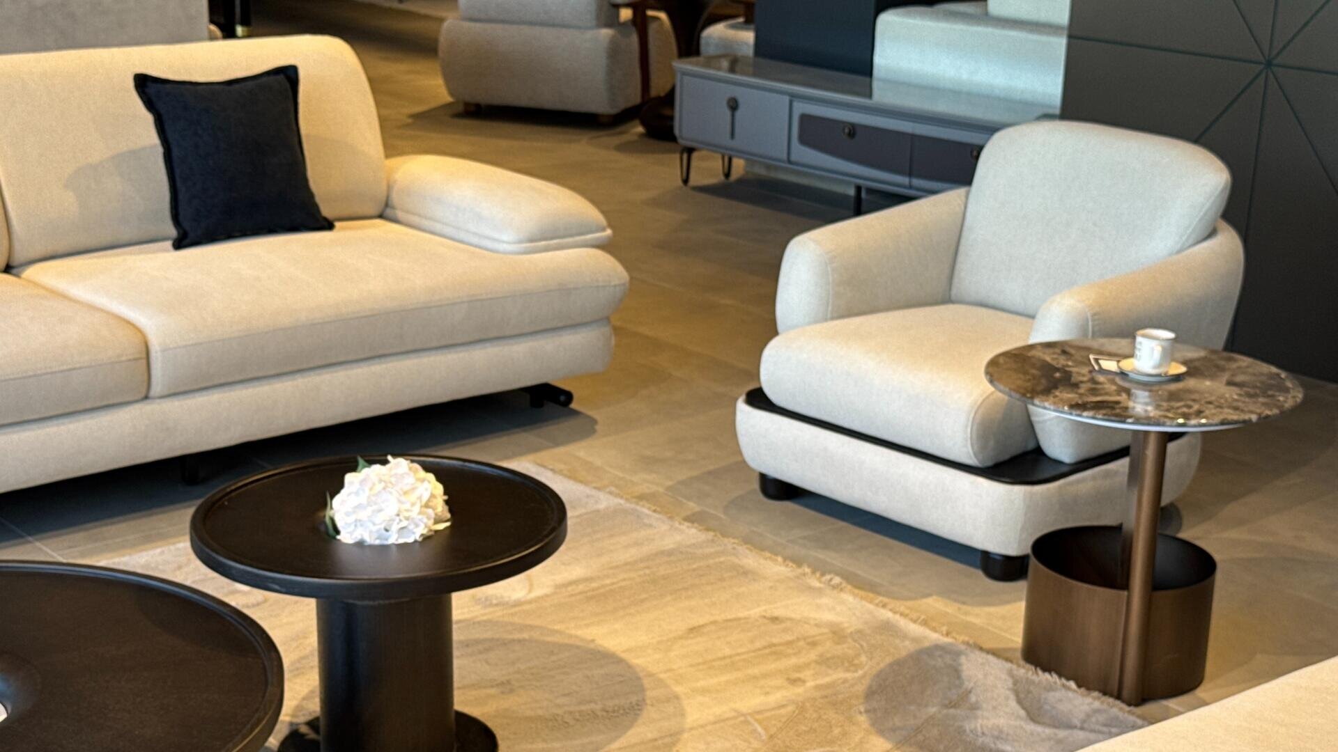 Ein modernes Wohnzimmer mit einem beigen Sofa und einem passenden Sessel, schwarzen und marmornen Beistelltischen, einem Blumenarrangement, einer Kaffeetasse und einem neutralen Teppich auf einem gefliesten Boden.