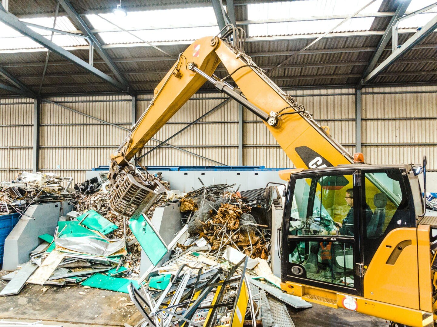 Ein gelber Bagger wühlt in einem großen Industrielager mit hoher, offener Decke in Stapeln von Metall und Schutt. Ein Arbeiter ist zu sehen, der die Maschine bedient.