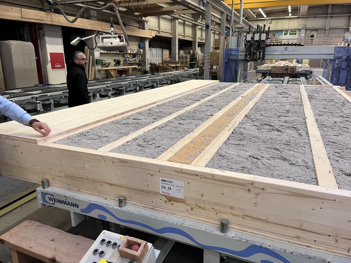 Ein großer Holzrahmen, gefüllt mit losem, grauem Isoliermaterial, steht in einer Fabrikumgebung. Links ist die Hand einer Person zu sehen, und eine weitere Person geht im Hintergrund neben Maschinen und Materialstapeln.