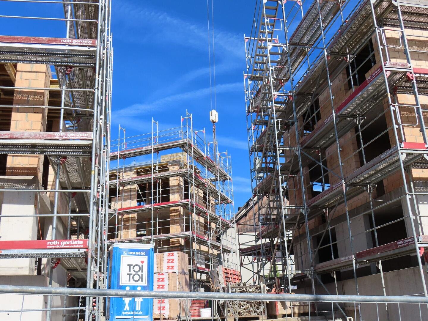 Eine Baustelle mit mehreren im Bau befindlichen Gebäuden, umgeben von Metallgerüsten. Eine blaue tragbare Toilette und Baumaterialien sind zu sehen, darüber ein klarer blauer Himmel.