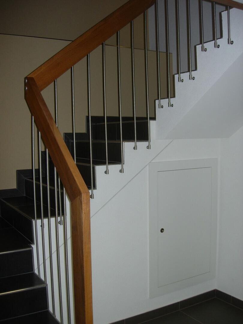 Un escalier intérieur moderne avec des marches noires, une rampe en métal et une main courante en bois. Sous l'escalier se trouve une petite porte blanche encastrée dans le mur.
