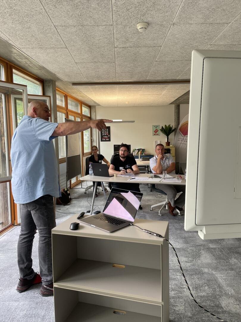 Ein Mann steht und zeigt auf einen Bildschirm, während er vor drei sitzenden Personen in einem modernen, hellen Konferenzraum mit Laptops und Papieren auf dem Tisch eine Präsentation hält.