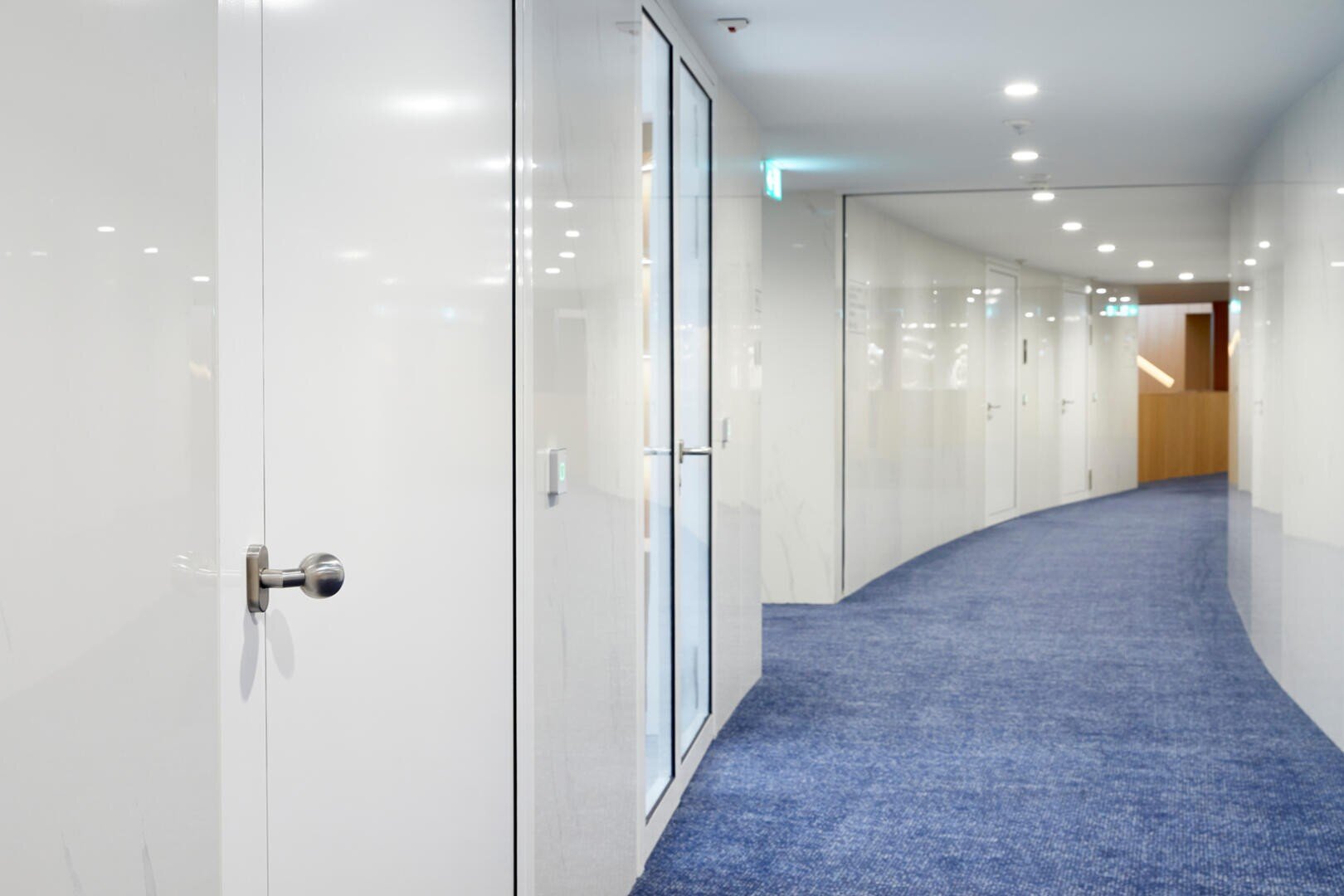 Un couloir moderne, brillamment éclairé, avec des murs blancs, des portes vitrées et une moquette bleue incurvée, qui mène à une autre zone au bout du couloir.