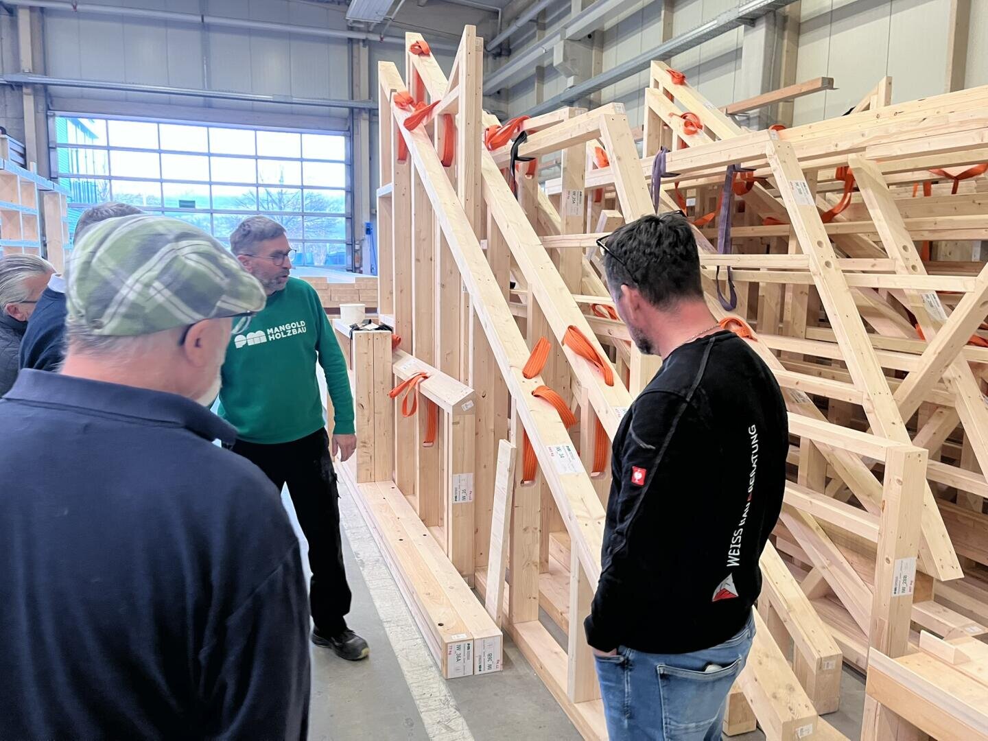 Drei Männer stehen in einer Werkstatt und betrachten große Holzkonstruktionen und Balken mit orangefarbenen Klammern. Ein Mann in einem grünen Hemd scheint etwas zu erklären, während die anderen zuhören. Im Hintergrund sind Regale und Fenster zu sehen.