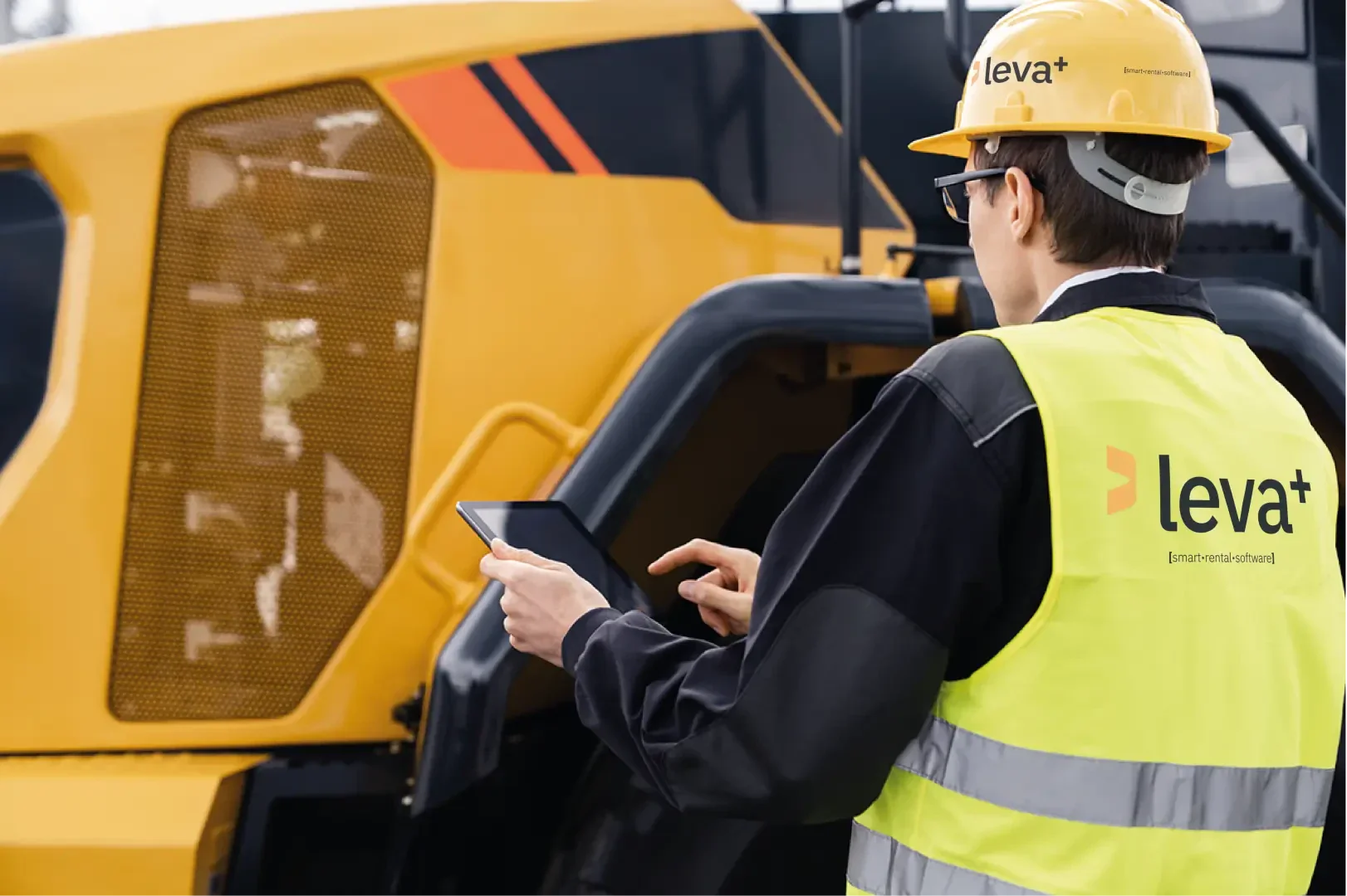 Ein Arbeiter, der einen gelben Schutzhelm und eine Warnweste mit der Aufschrift leva+ trägt, verwendet ein Tablet, während er neben einer großen gelben Industriemaschine steht.