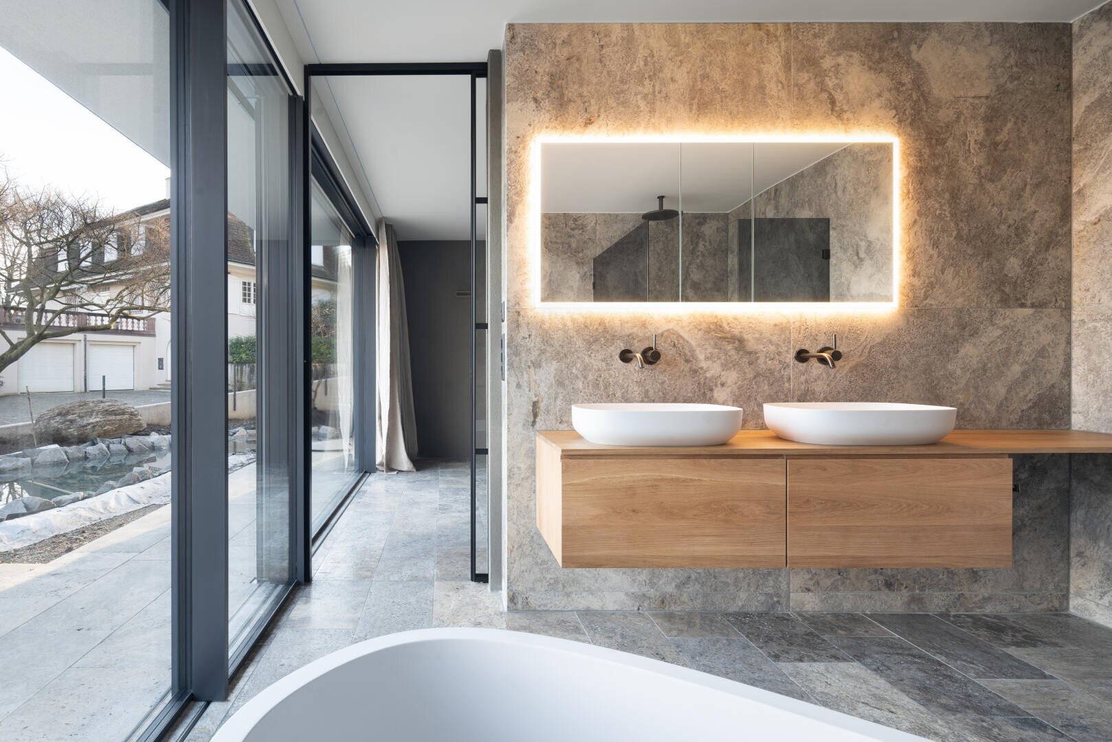 Modernes Badezimmer mit großen Fenstern, Steinwänden und -böden, einem schwebenden hölzernen Waschtisch mit zwei Gefäßwaschbecken, einem hinterleuchteten Spiegel und einer freistehenden Badewanne im Vordergrund. Der Raum ist von natürlichem Licht durchflutet.