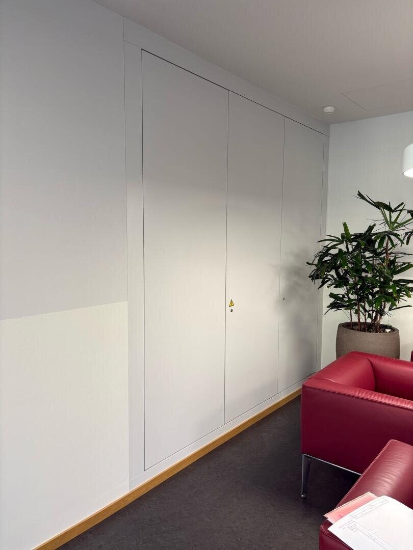 Ein hoher, weißer Einbauschrank mit dezenten Griffen steht vor einer hellen Wand. Eine große Topfpflanze und ein roter Sessel stehen in der Nähe. Auf der Armlehne des Sessels liegt ein Blatt Papier.