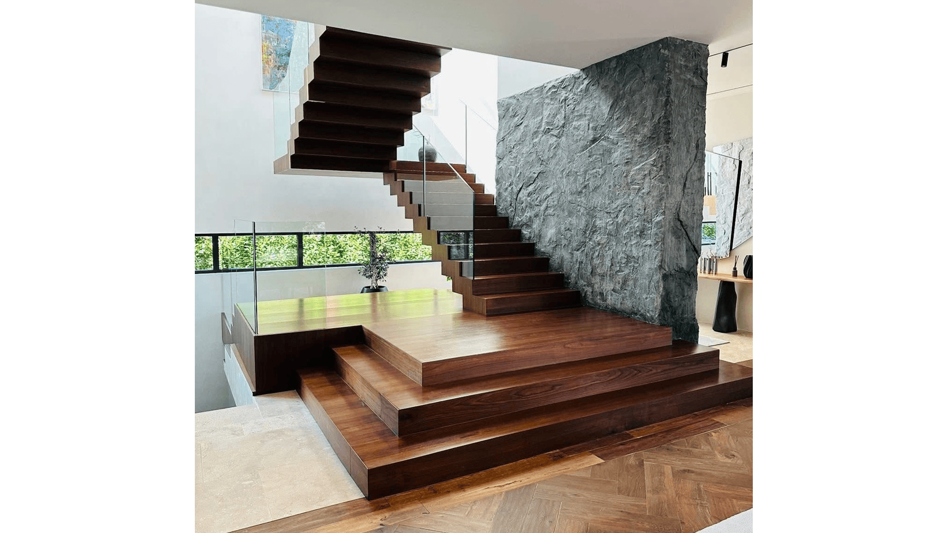 Eine moderne, offen gestaltete Treppe mit breiten, dunklen Holzstufen und Glasgeländer führt neben einer strukturierten grauen Steinwand in einem hellen, modernen Haus mit großen Fenstern und natürlichem Licht nach oben.