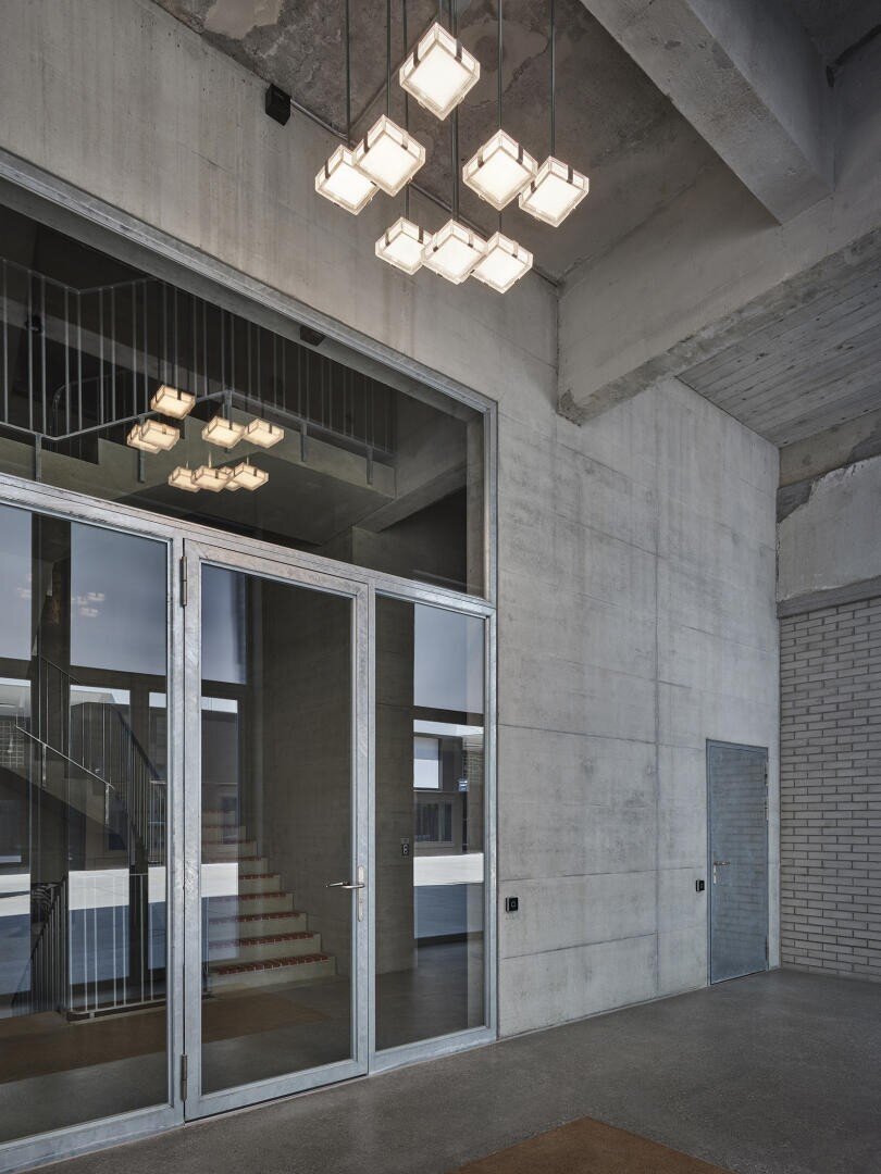 Ein modernes Interieur im Industriestil mit Betonwänden, einer Doppeltür aus Glas, einer geometrischen Leuchte und einer Metalltür. Eine Treppe und ein Geländer spiegeln sich im Glas. Der Raum wirkt sauber und minimalistisch.