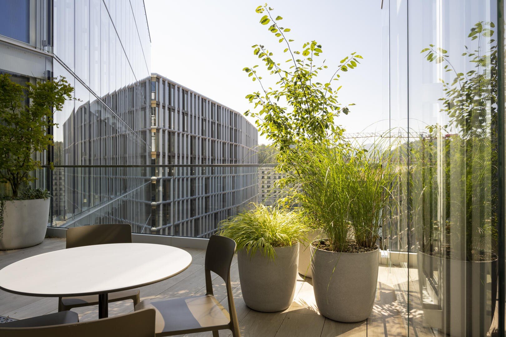 Sur un balcon moderne avec une balustrade en verre se trouvent une table ronde, des chaises et de grandes plantes en pot. En arrière-plan, on peut voir un bâtiment moderne à la façade grillagée en plein air.
