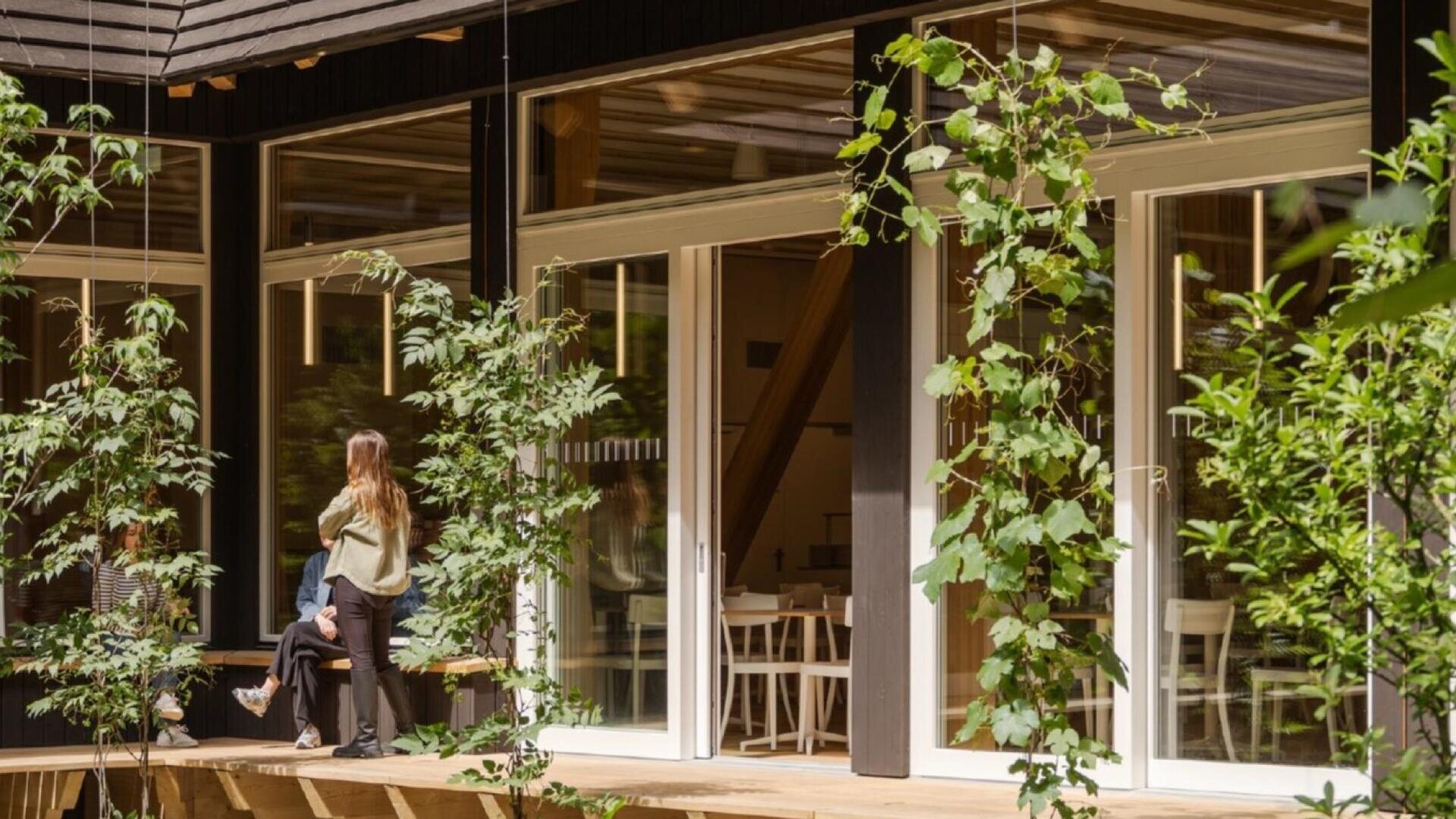 Eine Frau steht auf einer Holzterrasse vor einem modernen Gebäude mit großen Glastüren, umgeben von grünen Pflanzen und Bäumen in einem Innenhof.