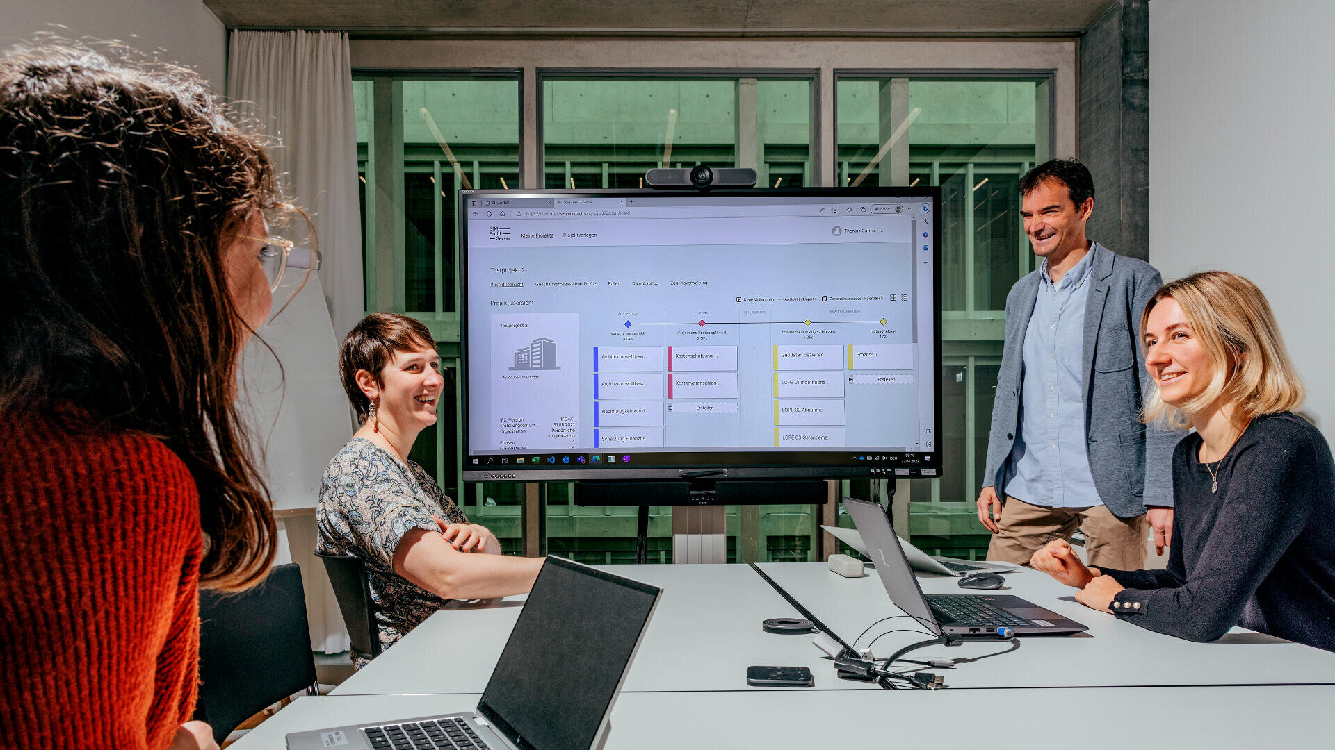 Vier Personen sitzen in einem Besprechungsraum und diskutieren über Inhalte, die auf einem großen Bildschirm angezeigt werden. Sie lächeln und sind engagiert, ihre Laptops liegen in einer modernen Büroumgebung auf dem Tisch.