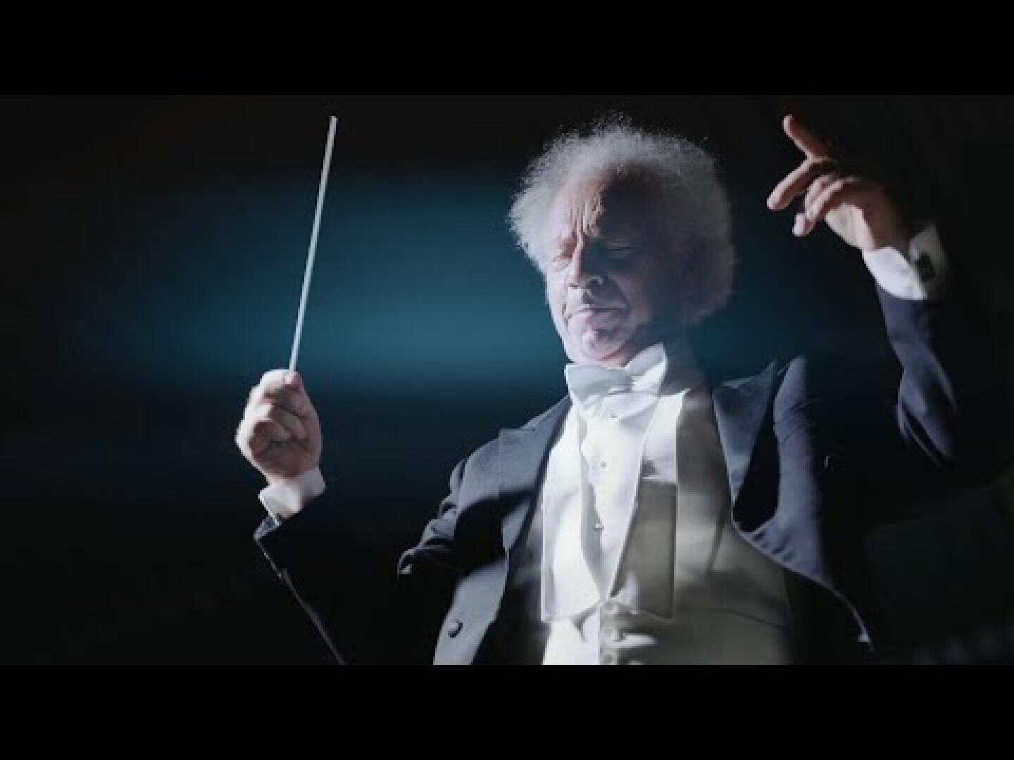 Un chef d'orchestre âgé en smoking tient une baguette et lève la main tout en dirigeant un orchestre éclairé par un éclairage dramatique de la scène.