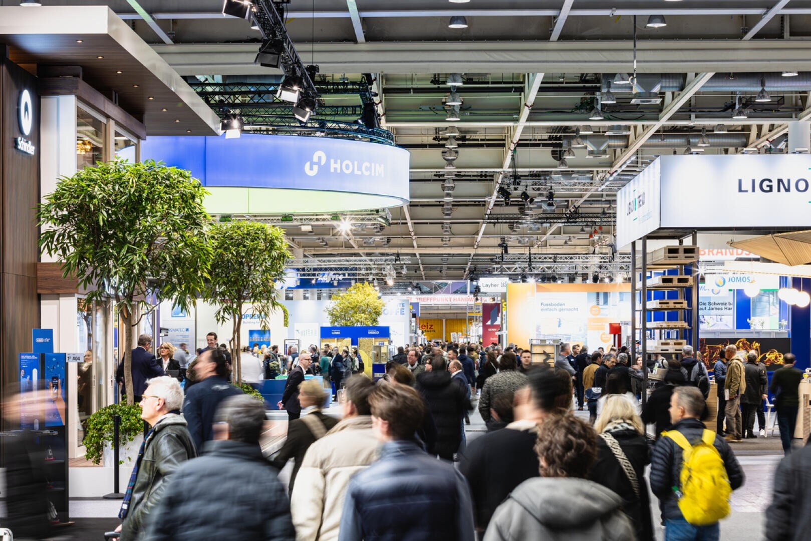 Eine geschäftige Indoor-Messe mit vielen Menschen, die zwischen den Messeständen umherlaufen, darunter auch Stände von Unternehmen wie Holcim. Helle Beleuchtung und Grünpflanzen schaffen eine lebendige Atmosphäre.