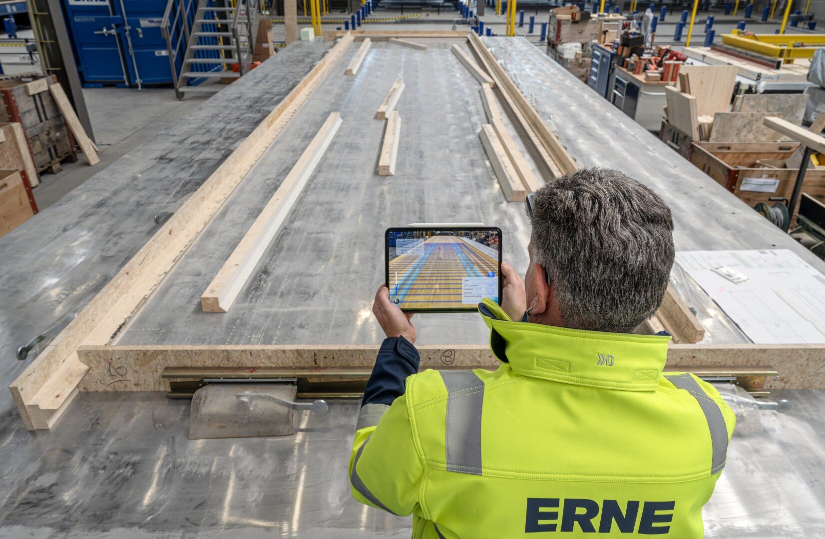 Ein Arbeiter in einer gelben ERNE-Jacke verwendet ein Tablet, um digitale Pläne über einer großen Metallfläche mit Holzbalken in einer Industriehalle zu betrachten.