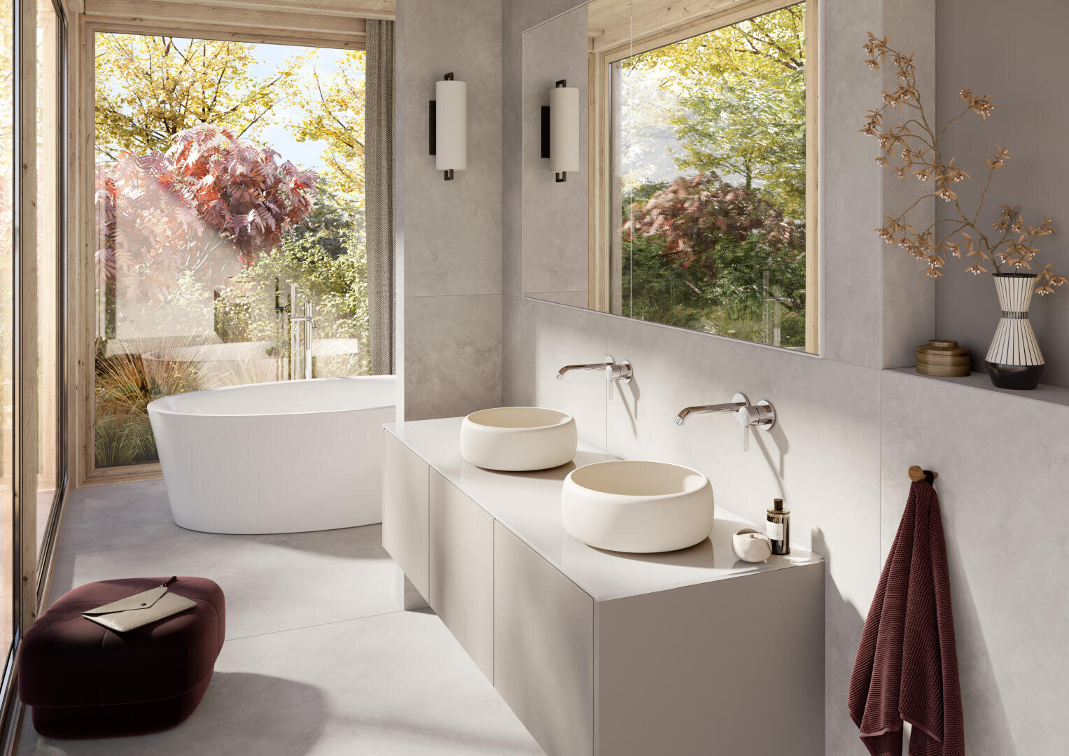 Salle de bain moderne avec une baignoire blanche indépendante près de grandes fenêtres, double lavabo sur une élégante table de toilette, robinetterie murale et arbres d'automne à l'extérieur. La décoration comprend une serviette marron, des vases en céramique et un tabouret moelleux.