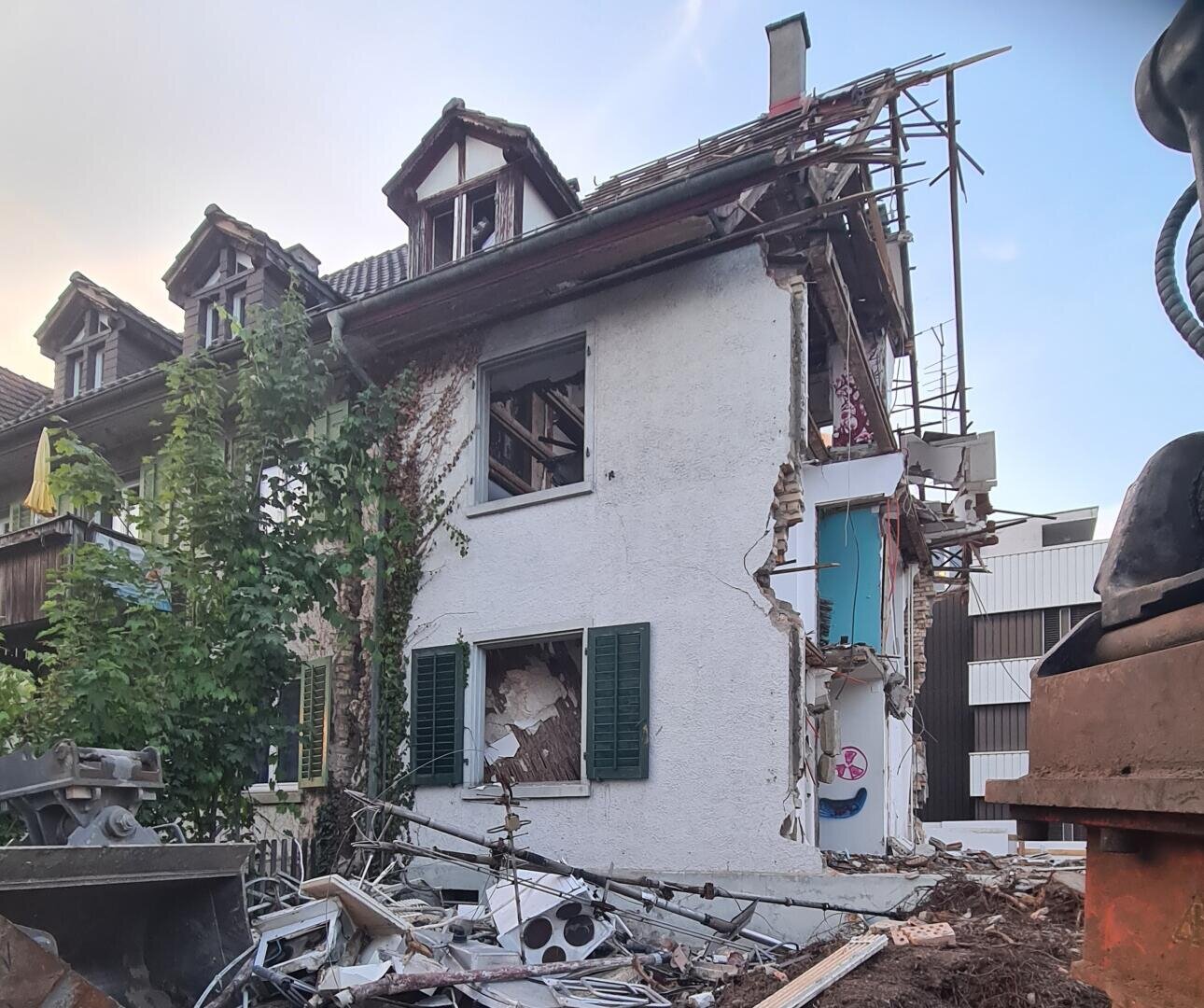 Ein teilweise zerstörtes weißes Haus mit freiliegenden Räumen und davor verstreuten Trümmern. Das Obergeschoss und das Dach sind beschädigt, und zerbrochene Fenster und Wände geben den Blick auf das Innere frei. In der Nähe ist Baumaterial zu sehen.