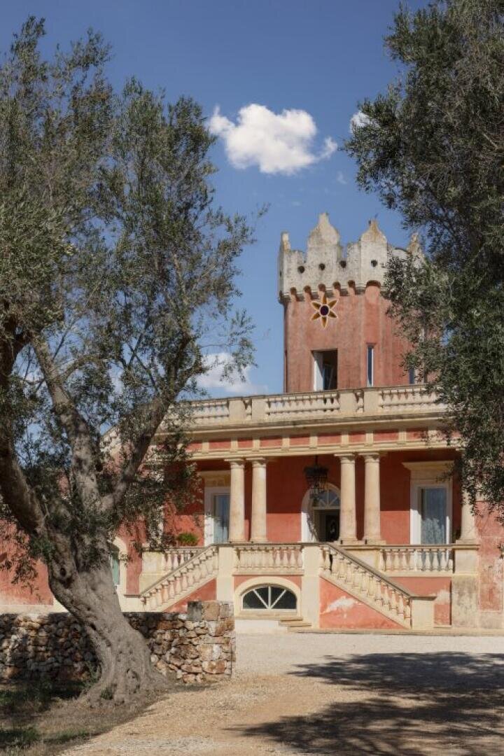 Une villa historique avec une façade rouge, des colonnes et une tour crénelée se dresse derrière des oliviers sous un ciel bleu avec un seul nuage blanc. Des marches en pierre mènent à l'entrée.