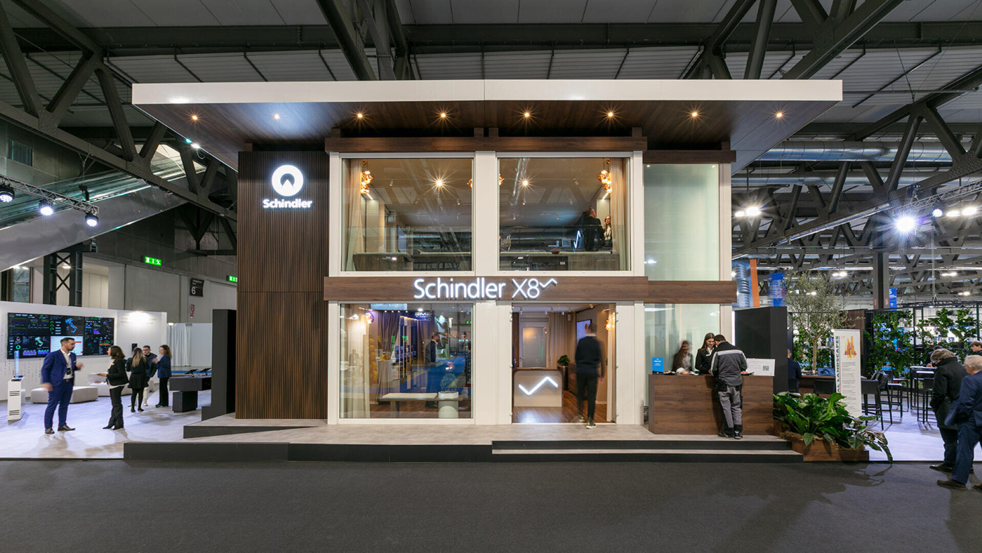 Zweistöckiger, moderner Messestand für Schindler mit großen Glasfenstern, Holzakzenten, beleuchteter Beschilderung und Menschen, die um den Stand herum in einem geräumigen Kongresszentrum interagieren.
