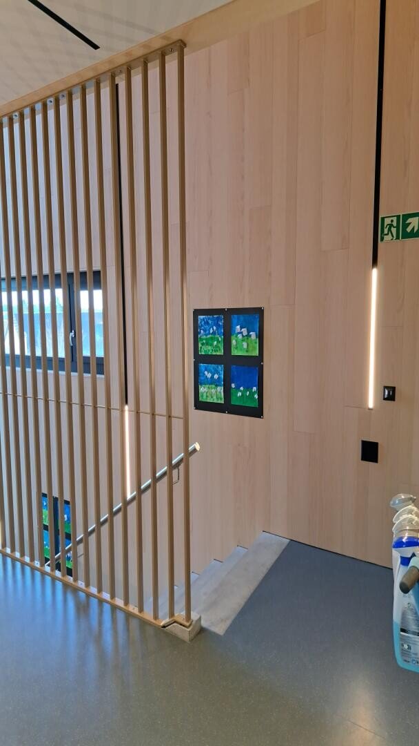 Ein modernes Innentreppenhaus mit Holzlatten, grauem Boden und Kunst an der Wand. Darüber befindet sich ein grünes Ausgangsschild, und in der unteren rechten Ecke stehen Reinigungsgeräte.