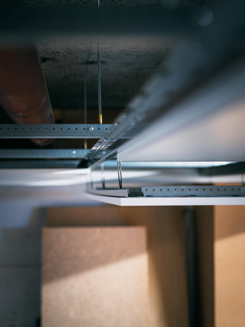 Gros plan sur une structure de plafond suspendue avec des suspensions et des panneaux métalliques, vue de dessous, avec des matériaux de construction et des tuyaux flous en arrière-plan.