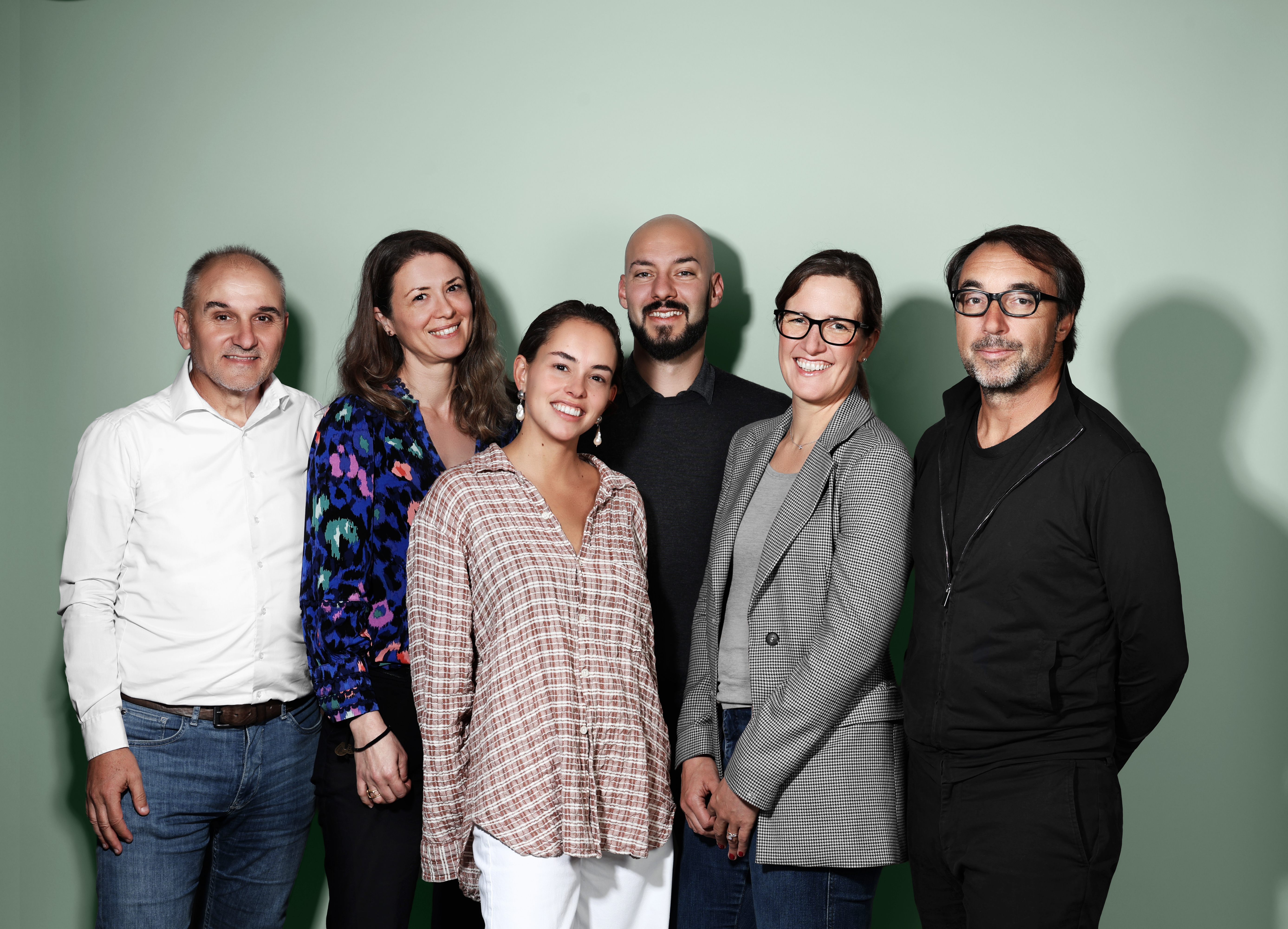 Six adultes se tiennent debout, souriants, devant un simple fond vert clair. Ils sont vêtus de vêtements décontractés et business casual et semblent détendus et amicaux.