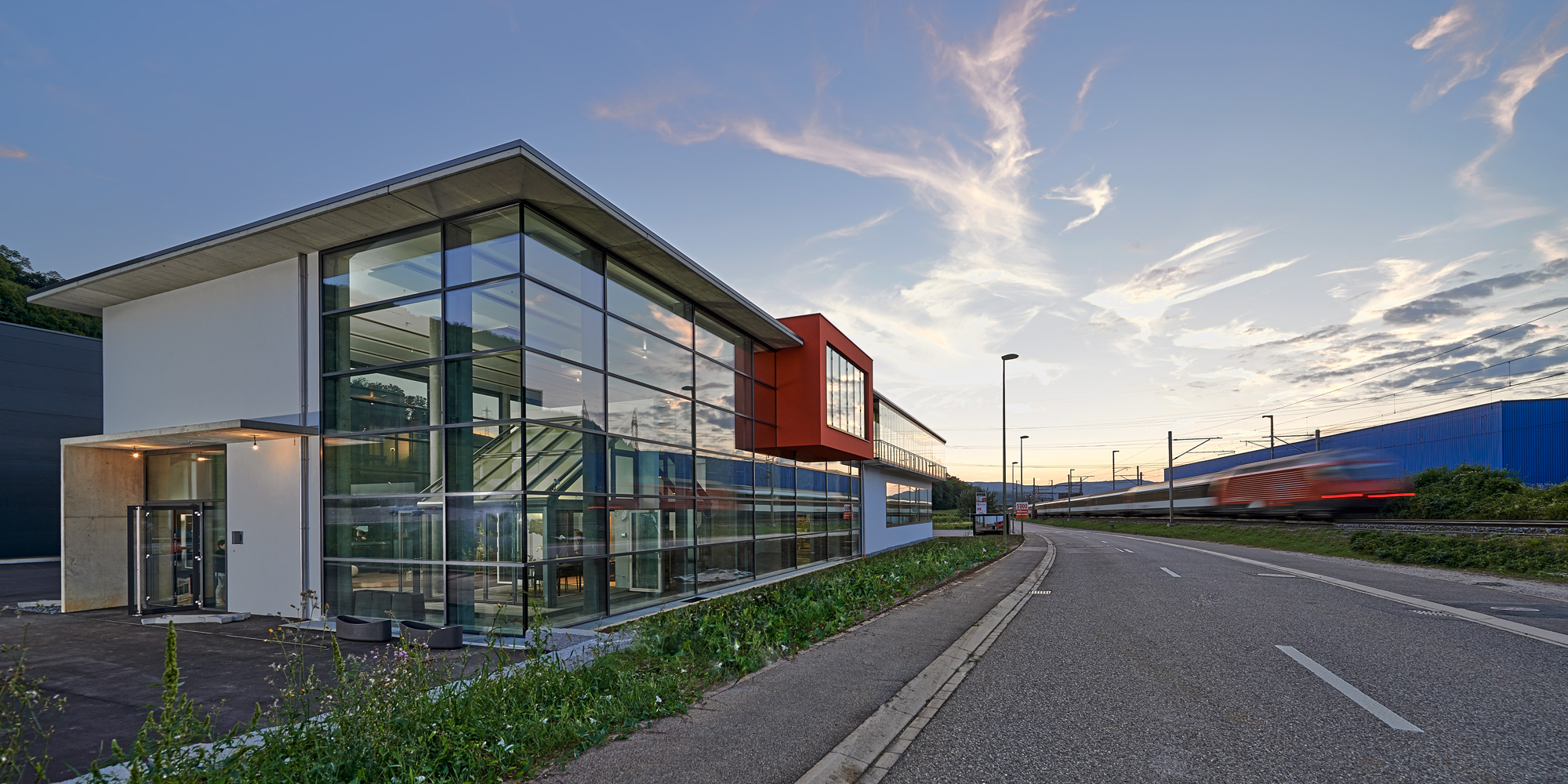 Ein modernes Gebäude aus Glas und Stahl mit scharfen Winkeln steht bei Sonnenuntergang neben einer leeren Straße, während auf den nahe gelegenen Gleisen ein verschwommener Zug vorbeifährt. Am blauen und orangefarbenen Himmel sind dünne Wolken zu sehen.