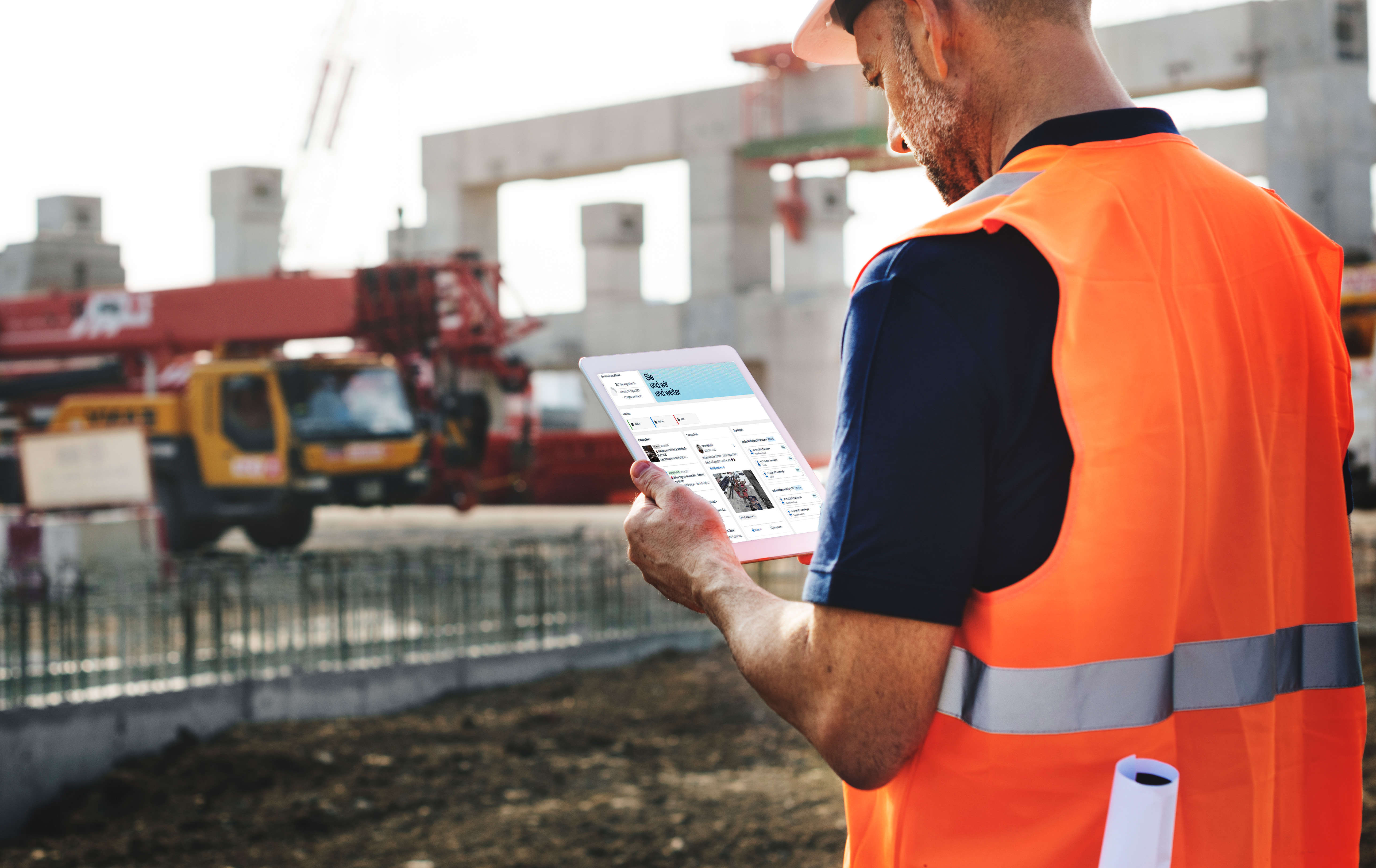 Ein Bauarbeiter mit orangefarbener Sicherheitsweste und Schutzhelm verwendet ein Tablet auf einer Baustelle mit Kränen und Gebäuderahmen im Hintergrund. Aufgerollte Baupläne sind unter einem Arm eingeklemmt.