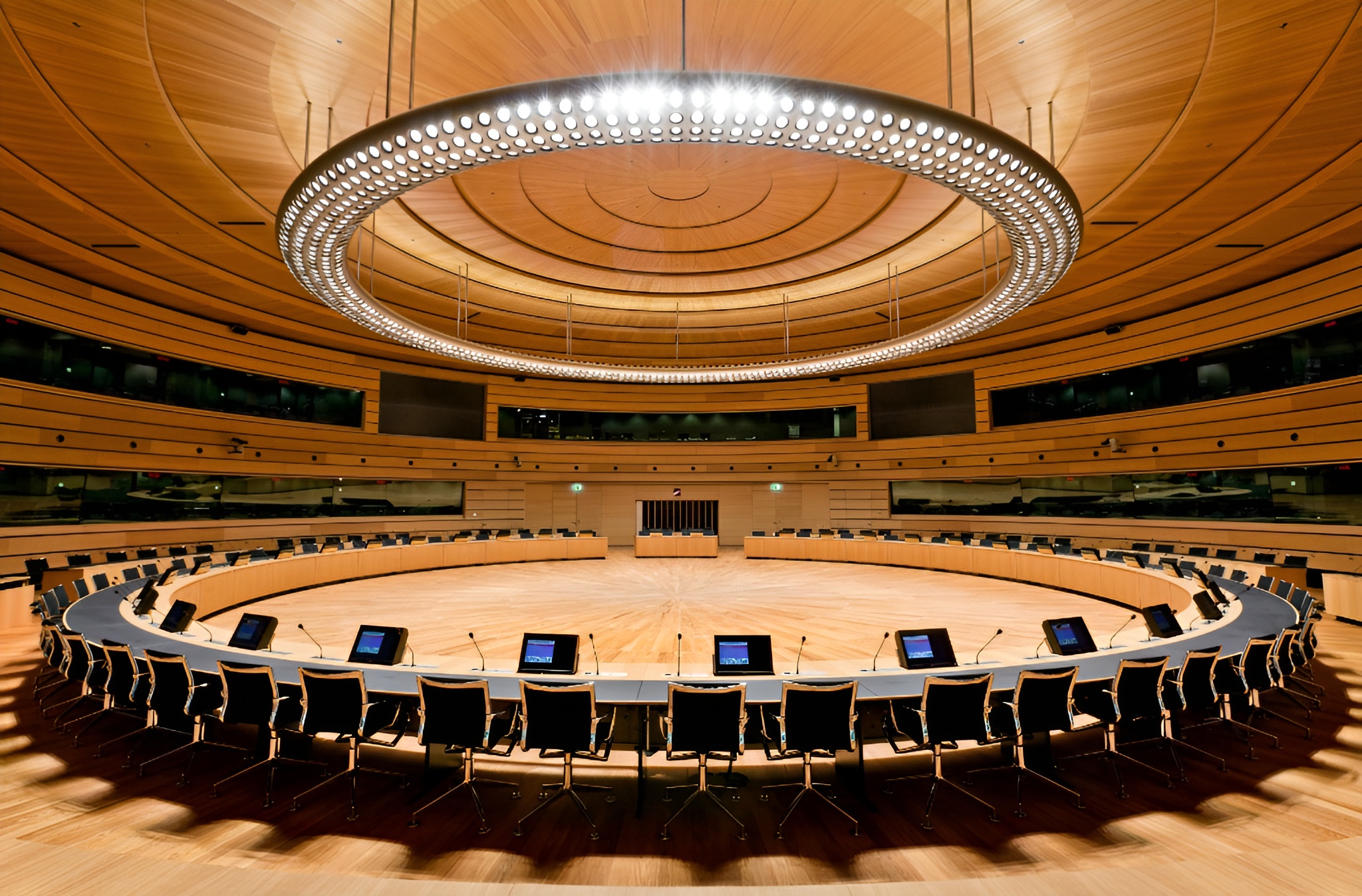 Une grande salle de conférence moderne avec une disposition circulaire de bureaux, de chaises noires, d'écrans d'ordinateur, et un luminaire rond au centre du plafond. La salle comporte des panneaux de bois et des sièges en gradins.