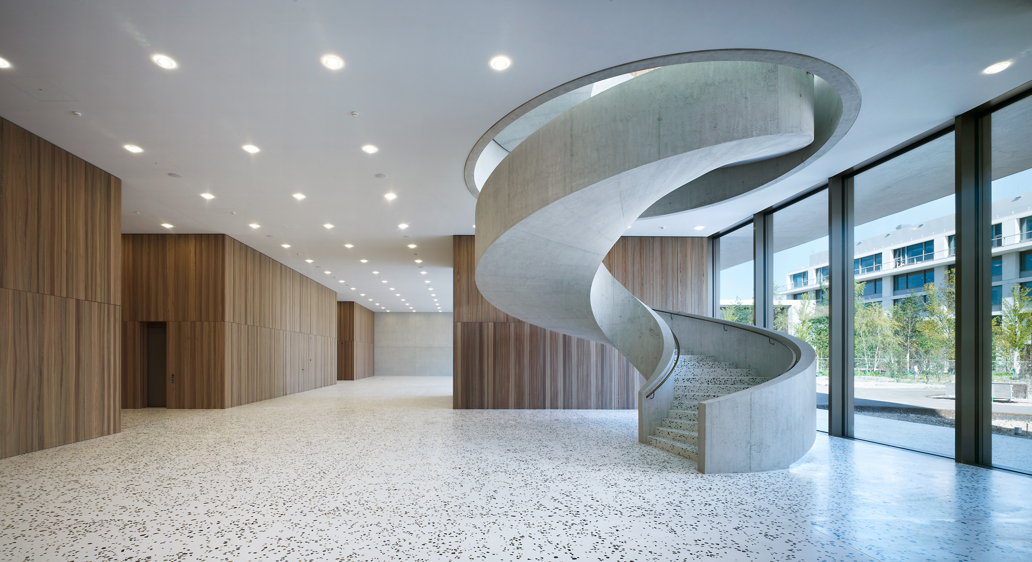 Hall d'entrée moderne avec des panneaux muraux en bois, un sol en terrazzo et un grand escalier en spirale en béton incurvé menant à une mezzanine. Des fenêtres allant du sol au plafond permettent de voir les arbres et les bâtiments à l'extérieur.