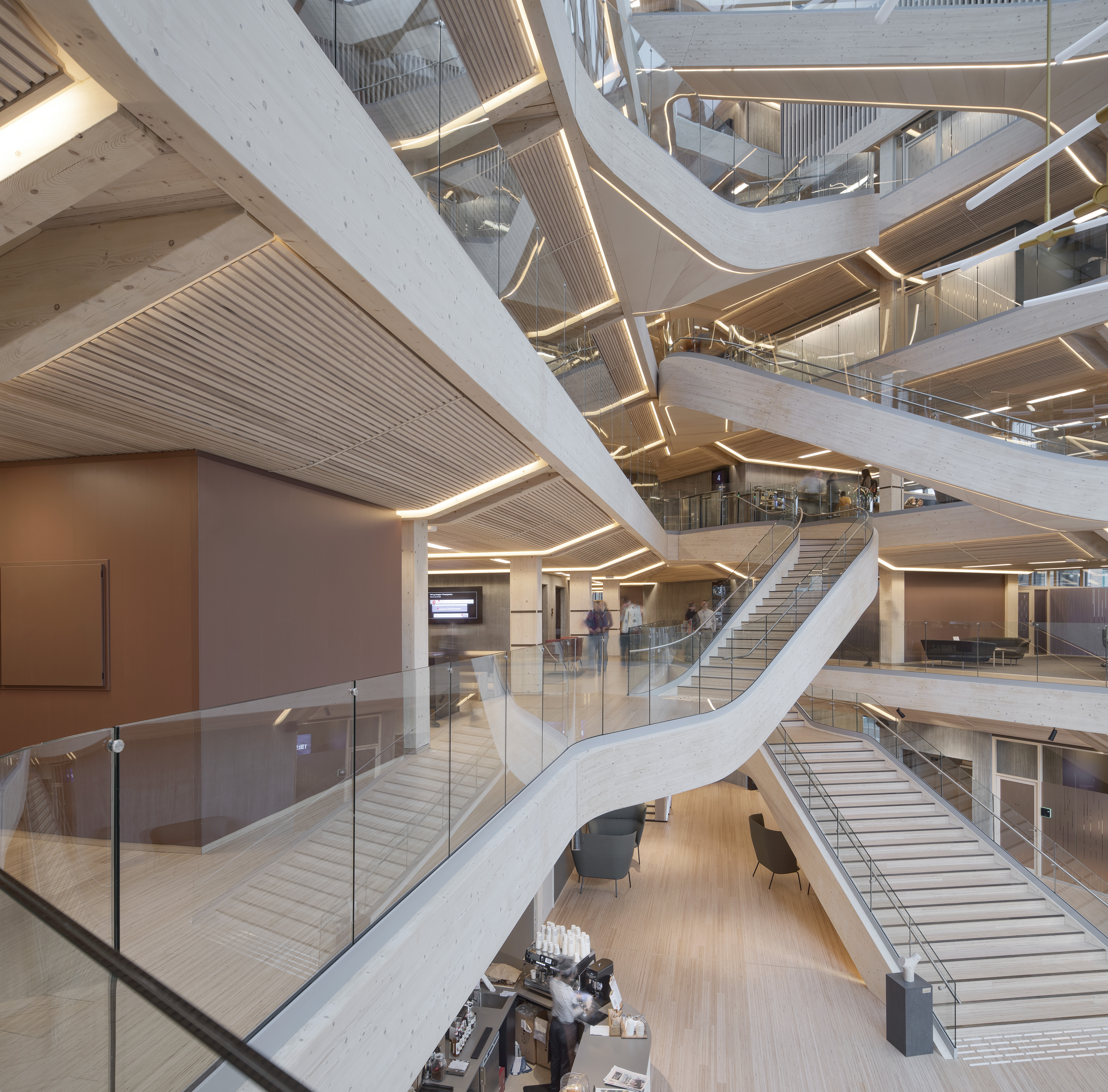Intérieur d'un bâtiment moderne à plusieurs niveaux, avec des escaliers en bois, des rampes en verre et un éclairage lumineux. Plusieurs personnes se promènent ou s'assoient dans l'atrium ouvert et spacieux, doté d'éléments de design contemporains.