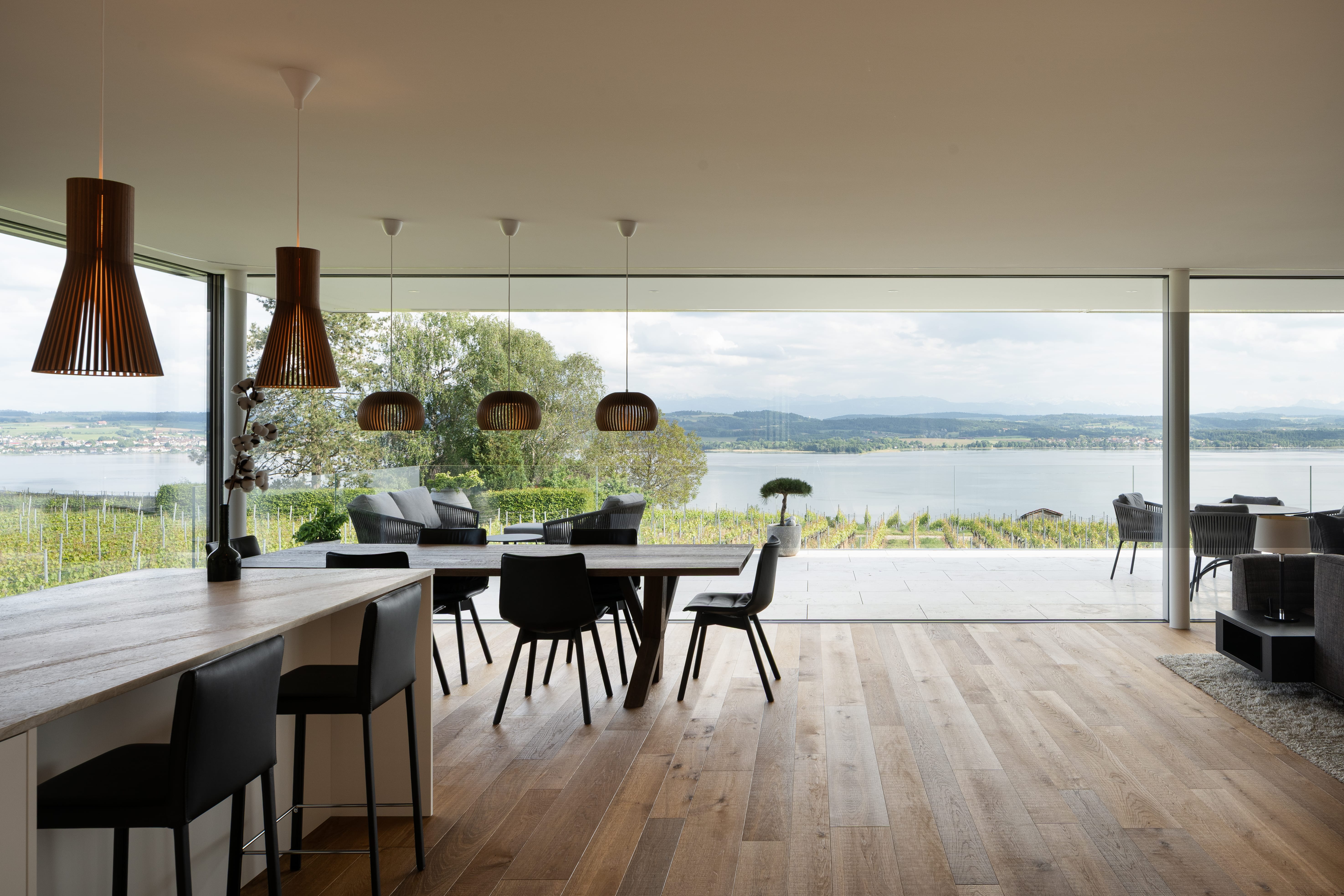 Salle à manger et salon modernes avec sols en bois, chaises noires et lampes suspendues, dotés de grandes baies vitrées offrant une vue panoramique sur un lac, de la verdure et des collines lointaines. Des sièges extérieurs sont visibles sur le patio.
