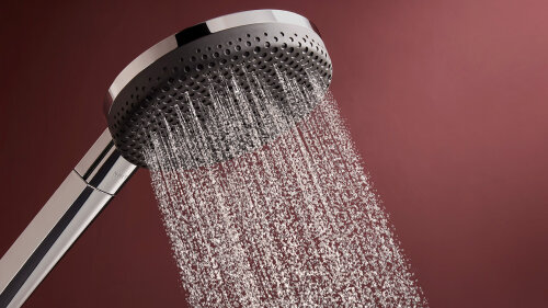 Eine Nahaufnahme einer modernen Handbrause, die Wasser versprüht, vor einem einfarbigen kastanienbraunen Hintergrund.