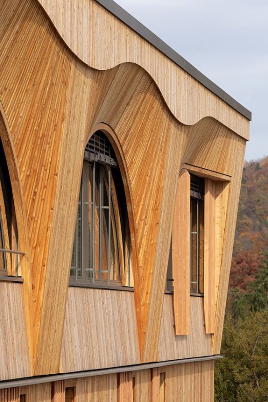 Nahaufnahme einer Gebäudefassade mit gewölbten Fenstern und komplizierten Holzverkleidungen mit geschwungenen geometrischen Mustern vor einem Hintergrund aus Bäumen und einem bewölkten Himmel.