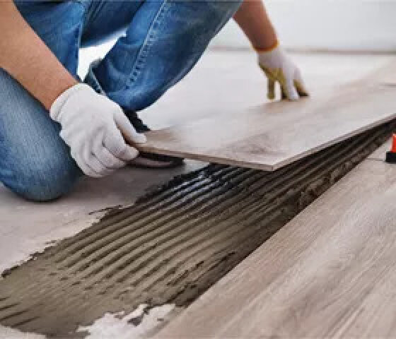 Une personne portant des gants blancs pose un parquet en bois en plaçant une lame sur la colle au sol.