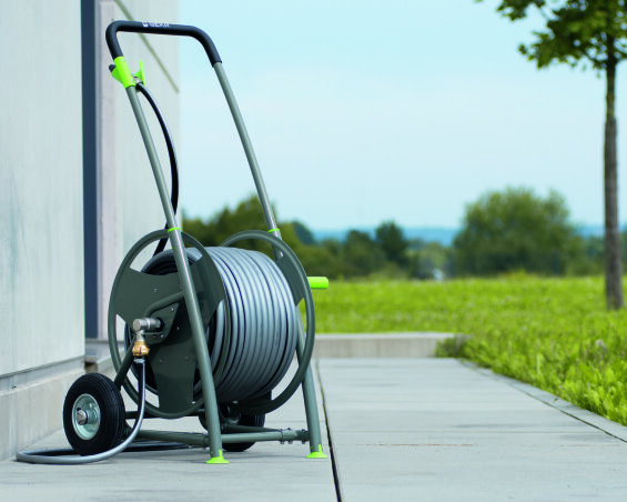 Ein Gartenschlauchwagen mit einem grauen Schlauch steht auf einem gepflasterten Gehweg neben einem Gebäude, im Hintergrund sind eine Wiese und Bäume zu sehen.