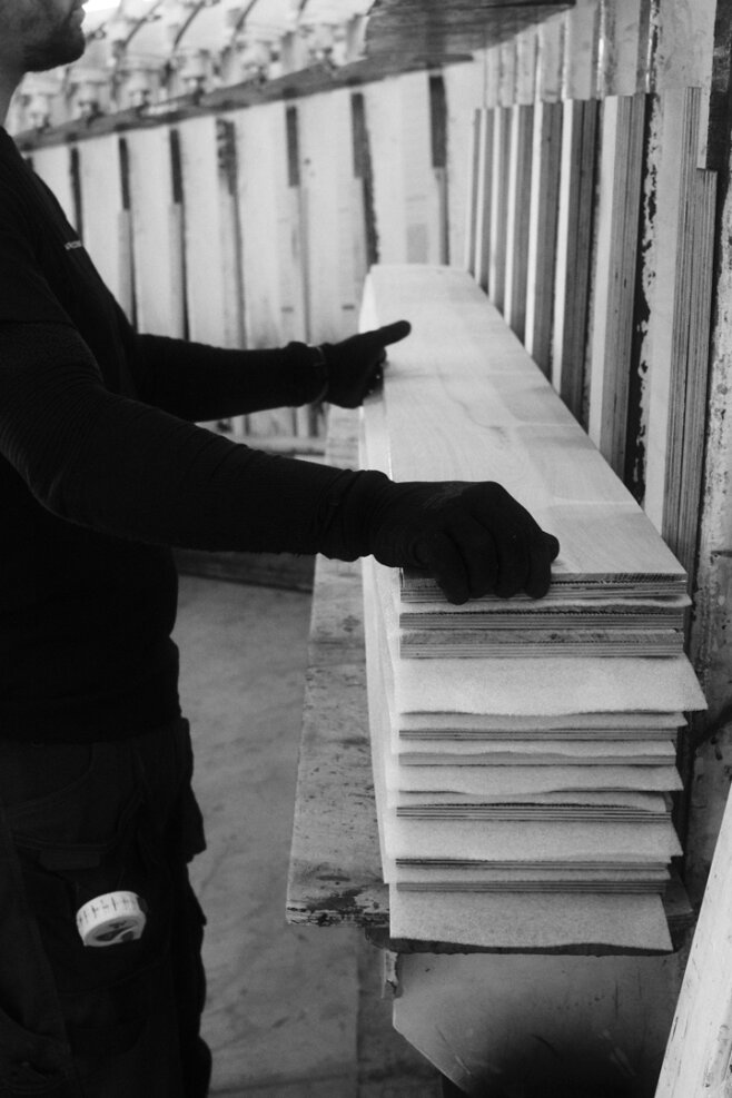 Eine Person mit Handschuhen ordnet einen Stapel Holzbretter oder -platten auf einem Arbeitstisch in einer Industrie- oder Werkstattumgebung. Die Szene ist in schwarz-weiß gehalten.