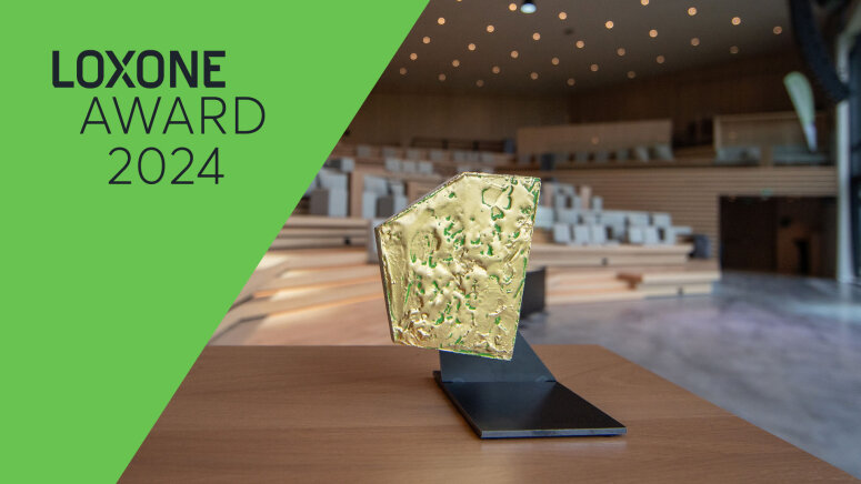 Eine goldfarbene Loxone Award-Trophäe steht auf einem schwarzen Ständer in einem modernen, leeren Auditorium. Die Worte LOXONE AWARD 2024 erscheinen auf einem grünen, diagonalen Banner auf der linken Seite des Bildes.