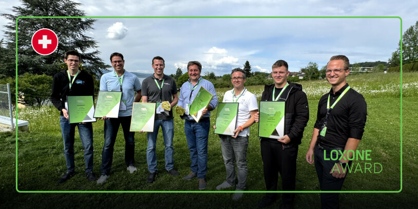 Sieben Männer stehen im Freien auf einer Wiese, lächeln und halten grüne Preisschilder in der Hand. Oben links ist das Symbol einer Schweizer Flagge zu sehen, und unten rechts steht "Loxone Award". Im Hintergrund sind Bäume und ein bewölkter Himmel zu sehen.