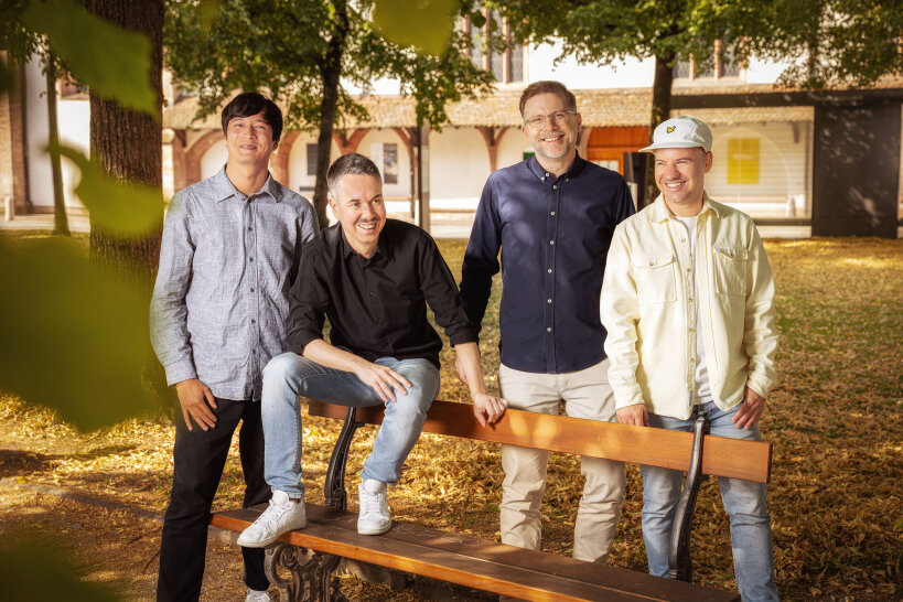 Vier Männer stehen und sitzen um eine Holzbank in einem Park mit Bäumen und herabgefallenen Blättern, lächelnd und leger in Hemden und Jacken gekleidet; im Hintergrund ist ein Gebäude zu sehen.